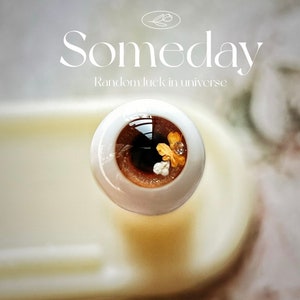 Puede incluir: Primer plano de un ojo de vidrio con iris marr&oacute;n y purpurina dorada. El ojo est&aacute; rodeado de peque&ntilde;as flores blancas y purpurina dorada. El texto "Someday Random luck in universe" es visible sobre el ojo.