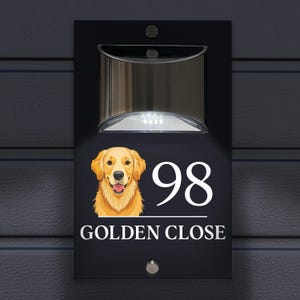Può includere: Una targa rettangolare nera per casa con una luce a energia solare sulla parte superiore, un'illustrazione grafica di un golden retriever, il numero 98 in bianco e il testo GOLDEN CLOSE stampato in lettere bianche nella parte inferiore.