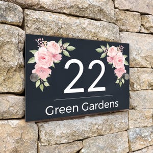 Peut inclure: Panneau d'adresse rectangulaire noir avec le numéro "22" en blanc et l'inscription "Green Gardens" en dessous. Décoré de compositions florales roses et de feuilles vertes. Le panneau est fixé sur un mur en pierre.