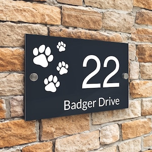 以下が含まれることがあります： 白い肉球の跡と数字の22、白い文字で「Badger Drive」と書かれた黒いアクリル製の家の番号札です。