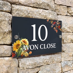 May include: A rectangular house sign with a dark grey background featuring the white number 10 and the text AVON CLOSE. The sign is decorated with yellow sunflowers and red rose hips, and it is mounted with two silver-coloured metal standoffs.