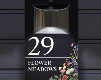Könnte beinhalten: Ein rechteckiges schwarzes Hausschild mit der Nummer 29 und dem Text FLOWER MEADOWS in weißer Schrift. Das Schild verfügt über eine integrierte solarbetriebene LED-Leuchte an der Oberseite und ein buntes Blumenmotiv auf der rechten Seite.