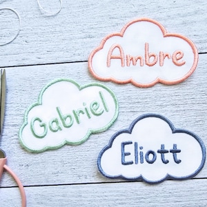 Può includere: Tre toppe ricamate a forma di nuvola con i nomi "Ambre", "Gabriel" e "Eliott". Ogni toppa ha un centro bianco, un bordo colorato e un nome. Forbici e filo sullo sfondo.
