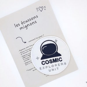 Può includere: Toppa termoadesiva bianca con un'illustrazione di astronauta blu e il testo "COSMIC EXPLORERS UNIT".