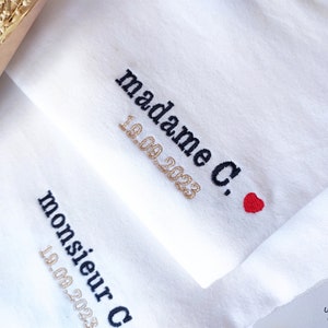 Op de afbeelding: Witte stof met zwarte borduurwerk dat "madame C." en "19.09.2023" in gouden draad leest. Een klein rood hart is geborduurd naast de tekst. De stof heeft ook zwarte borduurwerk dat "monsieur C." en "19.09.2023" in gouden draad leest.