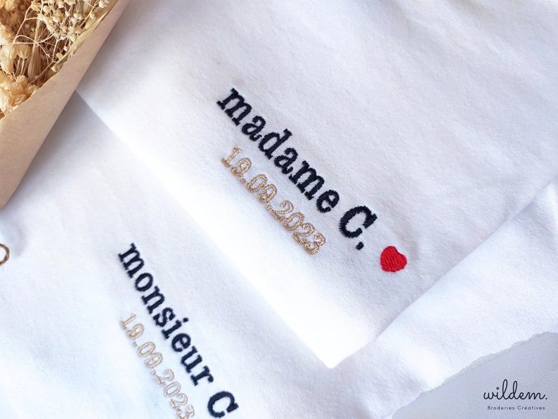 Op de afbeelding: Witte stof met zwarte borduurwerk dat "madame C." en "19.09.2023" in gouden draad leest. Een klein rood hart is geborduurd naast de tekst. De stof heeft ook zwarte borduurwerk dat "monsieur C." en "19.09.2023" in gouden draad leest.