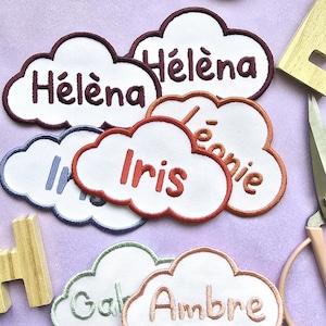 Könnte beinhalten: Gestickte wolkenförmige Aufnäher mit verschiedenen Namen. Namen sind unter anderem Héléna, Iris, Léonie, Ambre. Die Aufnäher haben weißen Stoff mit farbigen Rändern und Schrift. Eine Schere und Holzbuchstaben sind im Hintergrund.