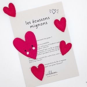 Peut inclure: Des écussons en forme de cœur en feutre rose avec des visages brodés. Les écussons sont sur une carte blanche avec le texte "les écussons mignons" et des instructions sur la façon de les repasser.