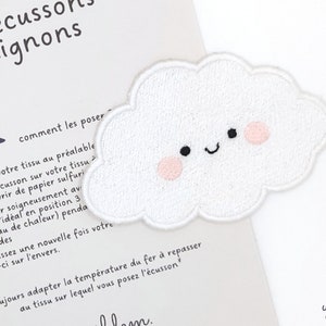 Puede incluir: Un parche bordado en forma de nube blanca con una cara sonriente y mejillas rosadas. El parche está sobre un fondo blanco con texto en francés que dice "les écussons mignons" y "comment les poser".