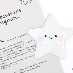Könnte beinhalten: Weißer, sternförmiger Bügel-Patch mit einem lächelnden Gesicht und rosa Wangen. Im Hintergrund ist der Text "les écussons mignons" und eine Anleitung zum Anbringen des Patches zu sehen.