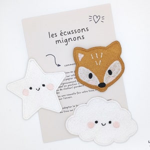 Könnte beinhalten: Drei gestickte Bügelbilder: ein weißer Stern, eine weiße Wolke und ein brauner Fuchs. Jedes Bügelbild hat ein lächelndes Gesicht mit rosa Wangen. Der Text "les écussons mignons" ist im Hintergrund sichtbar.