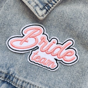 Peut inclure: Patch brodé rose et blanc avec le texte "Bride Team" sur un fond en jean.