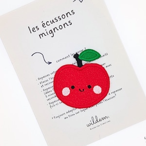 Peut inclure: Patch thermocollant en forme de pomme rouge en feutre avec un visage souriant et une tige verte. Le patch est sur un fond blanc avec des instructions sur la façon de le repasser. Le texte indique "les écussons mignons" et "wildem. Broderies Créatives".