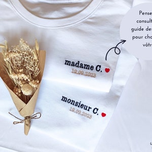 Op de afbeelding: Twee witte T-shirts met geborduurde tekst, "madame C. 18.09.2023" en "monsieur C. 18.09.2023", met een klein rood hartje naast elke naam. Een boeket gedroogde bloemen ligt op de T-shirts.
