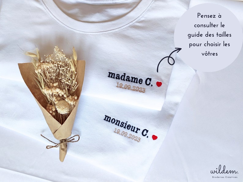 Op de afbeelding: Twee witte T-shirts met geborduurde tekst, "madame C. 18.09.2023" en "monsieur C. 18.09.2023", met een klein rood hartje naast elke naam. Een boeket gedroogde bloemen ligt op de T-shirts.