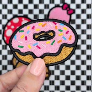 Può includere: Toppa ricamata a forma di ciambella rosa con codette colorate. La ciambella ha un contorno nero e un fondo glassato marrone.