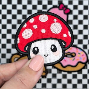 Può includere: Toppa ricamata raffigurante un fungo sorridente con cappello rosso e pois bianchi. Il fungo ha un viso bianco, occhi neri e un piccolo sorriso. La toppa è bordata di nero.