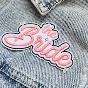 Peut inclure: Écusson brodé avec les mots "the Bride" en écriture cursive, soulignés en noir et rose. L'écusson est blanc avec des lettres roses et de petits accents d'étoiles. Il est fixé sur une veste en jean.