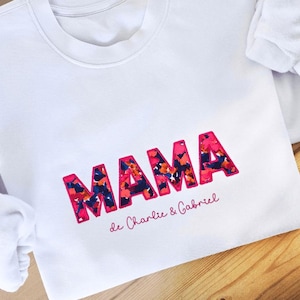 Peut inclure: Sweat-shirt blanc avec le mot "MAMA" en lettres roses soulignées avec un motif floral à l'intérieur. Le texte "de Charlie & Gabriel" est écrit en rose en dessous.