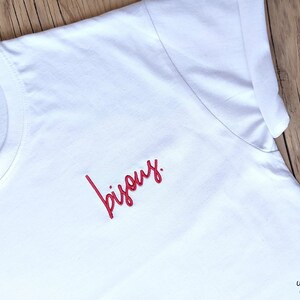 Op de afbeelding: Wit T-shirt met rode geborduurde tekst die "bisous" zegt.
