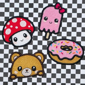 Peut inclure: Quatre patchs brodés thermocollants représentant un champignon rouge et blanc, une sucette glacée rose, un ours brun et un donut rose avec des pépites colorées.