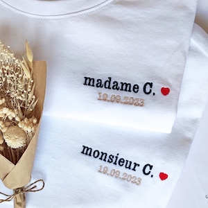 Peut inclure: Deux t-shirts blancs avec du texte brodé. Le t-shirt du haut dit "madame C." avec un cœur rouge et la date "19.09.2023". Le t-shirt du bas dit "monsieur C." avec un cœur rouge et la date "19.09.2023".