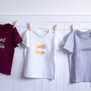 Op de afbeelding: Drie T-shirts hangen aan een waslijn. Het shirt links is bordeauxrood met witte borduurwerk dat "enfant terrible" zegt. Het middelste shirt is wit met gouden borduurwerk dat "petit loup" zegt. Het shirt rechts is grijs met zwarte borduurwerk dat "canaille" zegt.