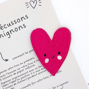 Può includere: Toppa a forma di cuore in feltro rosa con un viso sorridente e due guance rosa. La toppa è su uno sfondo bianco con istruzioni su come stirare la toppa.
