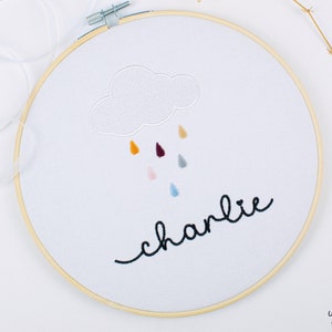 Peut inclure: Un cercle de broderie blanc avec un nuage blanc et des gouttes de pluie colorées brodées dessus. Le mot "charlie" est brodé en cursive noire sous le nuage.