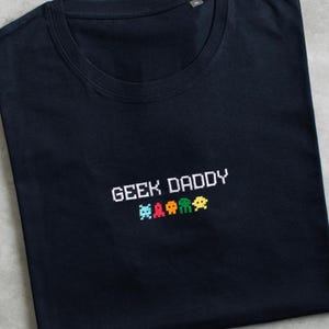 Może przedstawiać: Czarny T-shirt z haftem "GEEK DADDY" w kolorze białym. Pięć pikselowych kosmitów jest wyszytych pod tekstem w różnych kolorach.