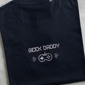 Può includere: Una maglietta blu scuro con testo ricamato bianco che recita "Geek Daddy" e una grafica di un controller per videogiochi pixelato.