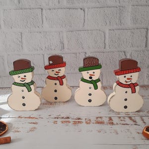 Könnte beinhalten: Vier hölzerne Schneemann-Figuren mit bemalten Details. Jeder Schneemann trägt einen braunen Hut, einen roten Schal und einen grünen Schal. Die Schneemänner stehen auf einer weißen Holzoberfläche.