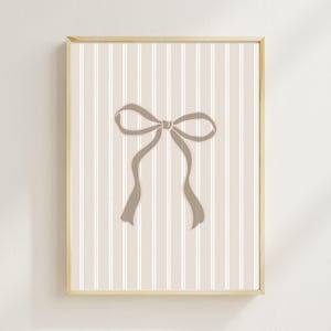 Puede incluir: Una impresión artística enmarcada con una ilustración de lazo color topo sobre un fondo a rayas. El lazo es de color topo apagado, y el fondo tiene rayas verticales en beige y blanco. El marco es de color madera clara.