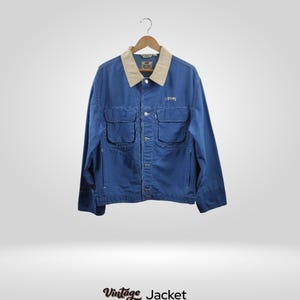Peut inclure: Veste vintage bleue avec un col beige et quatre poches avant. La veste est suspendue à un cintre en bois. Le mot "LEVIS" est brodé sur la poitrine. Le texte "Vintage Jacket" est en bas.
