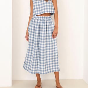 May include: A two-piece outfit featuring a blue and white gingham pattern. The set includes a sleeveless crop top and a midi-length skirt. The outfit is made of a lightweight fabric, perfect for warm weather. The model is wearing brown sandals.