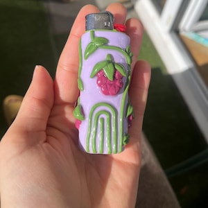 Peut inclure: Un briquet lilas avec un motif décoratif de vignes vertes, de feuilles et d'une framboise. Le briquet a un dessus argenté et un bouton rouge. Le design est peint à la main et ajoute une touche unique au briquet.