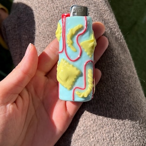 Peut inclure: Un briquet bleu clair avec un motif sur le thème des pâtes. Le briquet présente des formes de pâtes jaunes soulignées de lignes rouges. Le briquet est tenu dans une main, avec un fond marron.