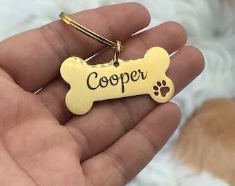 May include: Gold bone-shaped dog tag with the name "Cooper" engraved on it. The tag has a paw print cutout and a metal ring.