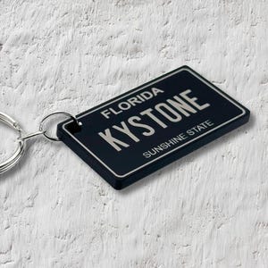 May include: Black keychain with a silver metal ring. The keychain features a black rectangular tag with the text "FLORIDA KYSTONE SUNSHINE STATE" in silver lettering.