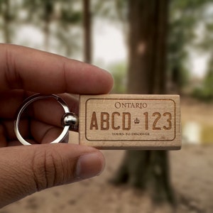 May include: Wooden keychain with an engraved Ontario license plate design. The plate reads "Ontario" at the top, with "ABCD 123" and "Yours to Discover" below. The keychain has a silver metal ring.