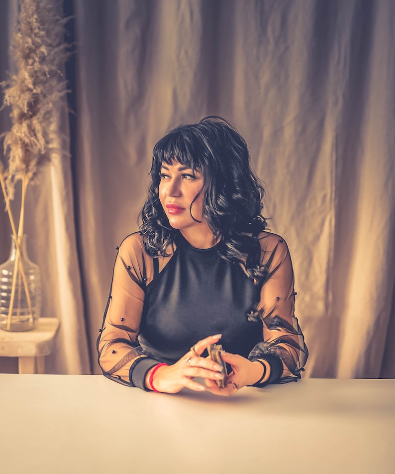 Peut inclure: Une femme aux cheveux fonc&eacute;s portant un haut noir transparent &agrave; pois noirs est assise &agrave; une table blanche, tenant un jeu de cartes dans ses mains.