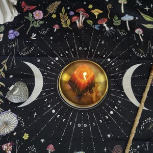 Peut inclure: Une tapisserie noire avec un motif de lune et d'&eacute;toiles blanches. Une bougie allum&eacute;e est plac&eacute;e dans un plat dor&eacute; au centre de la tapisserie. La tapisserie est entour&eacute;e d'une vari&eacute;t&eacute; de petits champignons color&eacute;s.