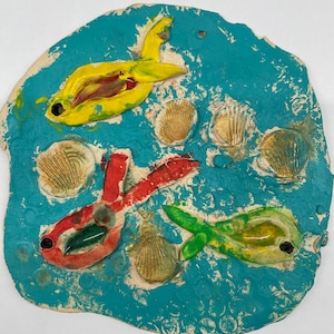 May include: A blue clay sculpture of three fish and seashells. The fish are yellow, red, and green with black eyes. The seashells are brown and gold.