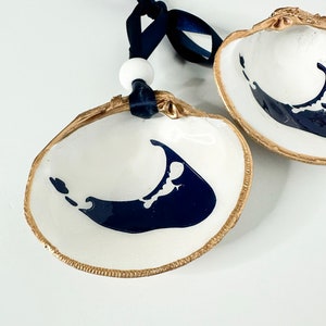 May include: Two white seashells with gold trim and a blue outline of an island. The shells are hanging from a blue ribbon with a white bead.