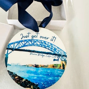 May include: A round, white ornament with a watercolor painting of the Bourne Bridge in Cape Cod, Massachusetts. The bridge is painted in shades of blue, and the water is a light blue. The text "Just get over it!" is written in black cursive above the bridge, and the text "Bourne Bridge, Cape Cod" is written in black cursive below the bridge.