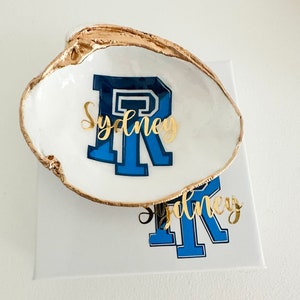 Op de afbeelding: Een wit schelpen sieradenschaaltje met een goudkleurige rand. Het schaaltje heeft de blauw-witte letter "R" met de naam "Sydney" in gouden script. Het schaaltje staat op een wit oppervlak.