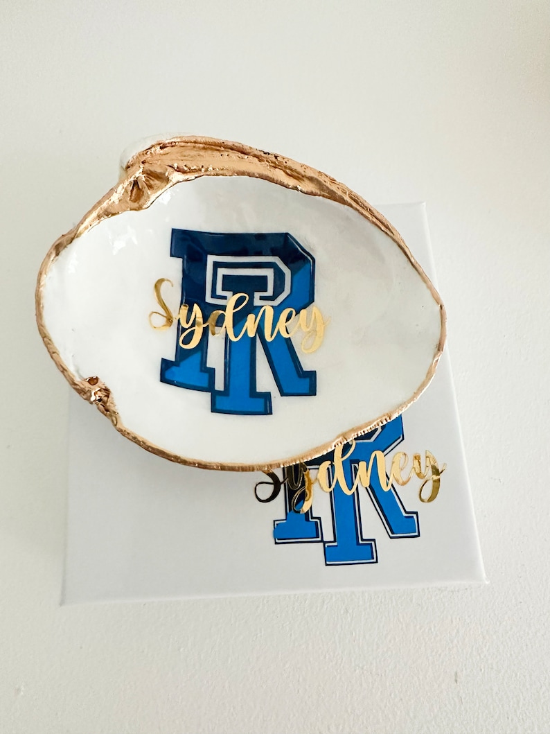 Op de afbeelding: Een wit schelpen sieradenschaaltje met een goudkleurige rand. Het schaaltje heeft de blauw-witte letter "R" met de naam "Sydney" in gouden script. Het schaaltje staat op een wit oppervlak.
