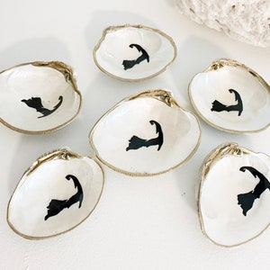 May include: Six white clam shells with gold trim, each painted with a black silhouette of Cape Cod. The shells are arranged on a white surface.