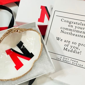 Op de afbeelding: Een witte oesterschelp schaal met een gouden rand, met een rood en zwart "N" logo, gepresenteerd in een witte doos. De afbeelding bevat een kaart met tekst die iemand feliciteert met zijn toewijding aan Northeastern.