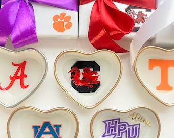 Könnte beinhalten: Herzförmige Schmuckschalen mit goldenen Rändern, jede mit einem anderen College-Logo. Weiße Geschenkboxen mit bunten Bändern und Logos befinden sich im Hintergrund. Die Logos enthalten die Buchstaben "A", "T", "HPU" und "AU" sowie einen Pfotenabdruck.